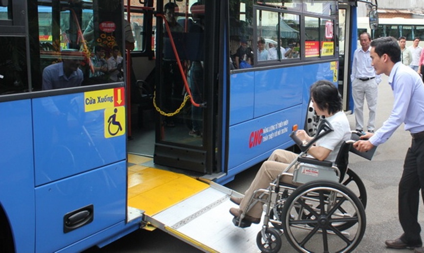 A women using a wheelchair enters a bus on a ramp and a companion pushes her wheelchair.