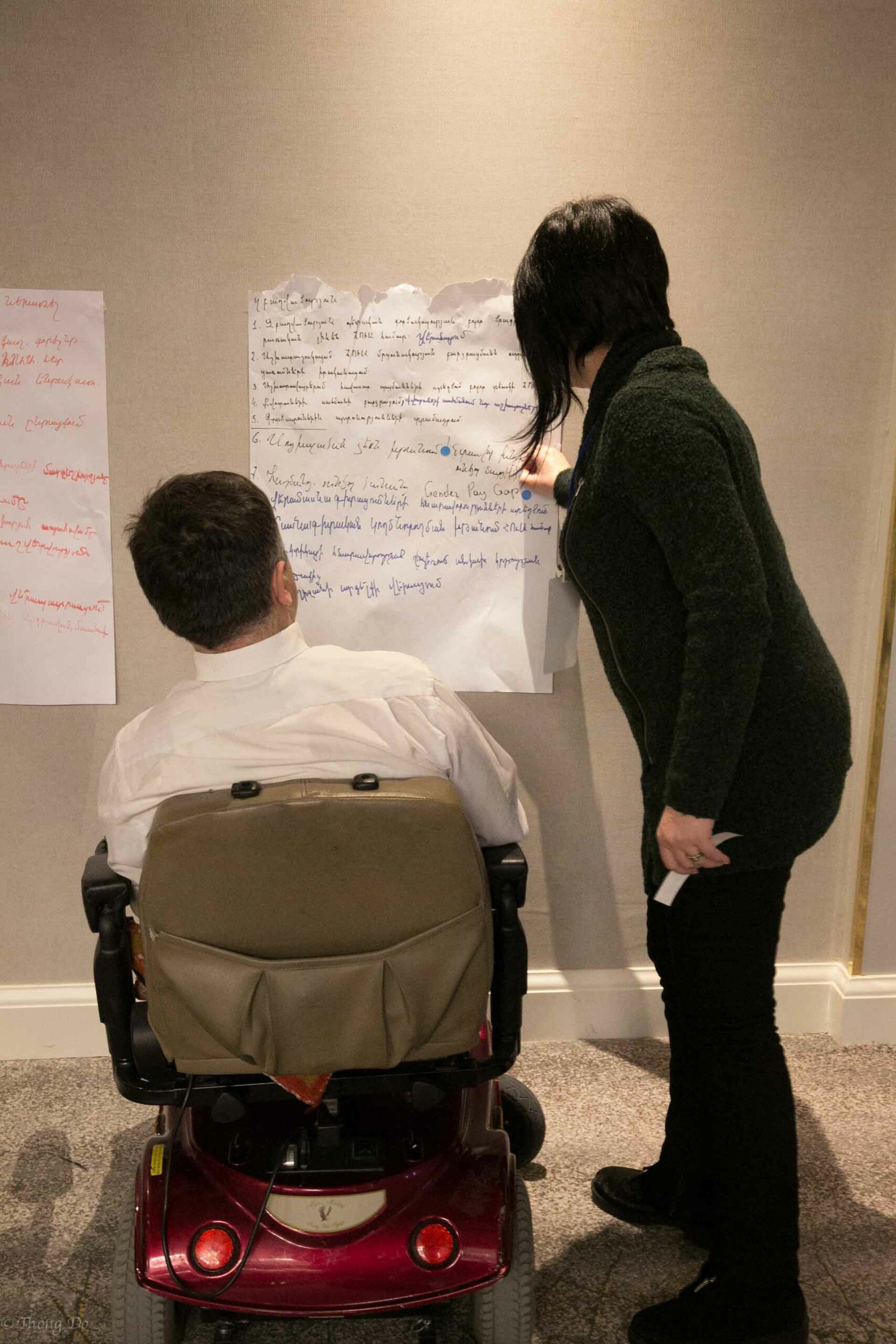 A man using a wheelchair and a woman standing next to him look at a flip chart paper on a wall.