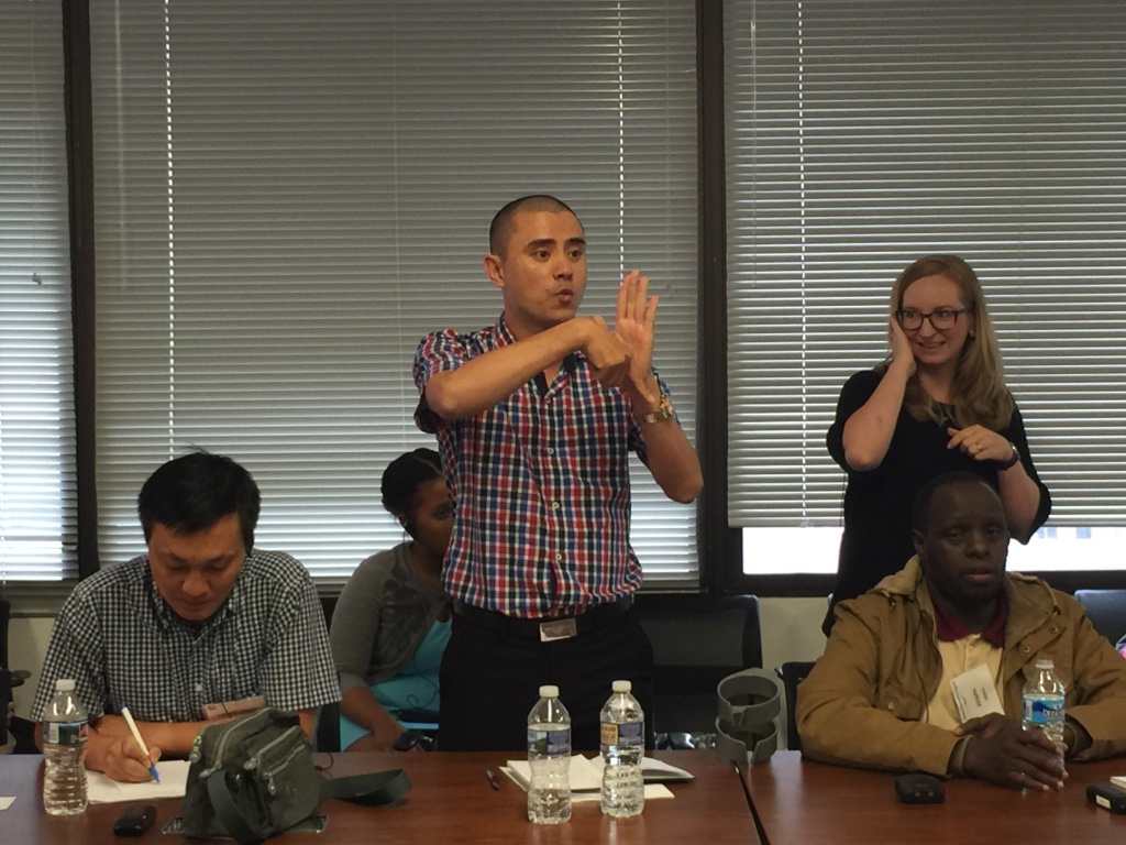 A Vietnamese man stands at a conference table speaking in Sign Language to someone who is out of the picture. He is surrounded by four seated people, one of whom is taking notes.