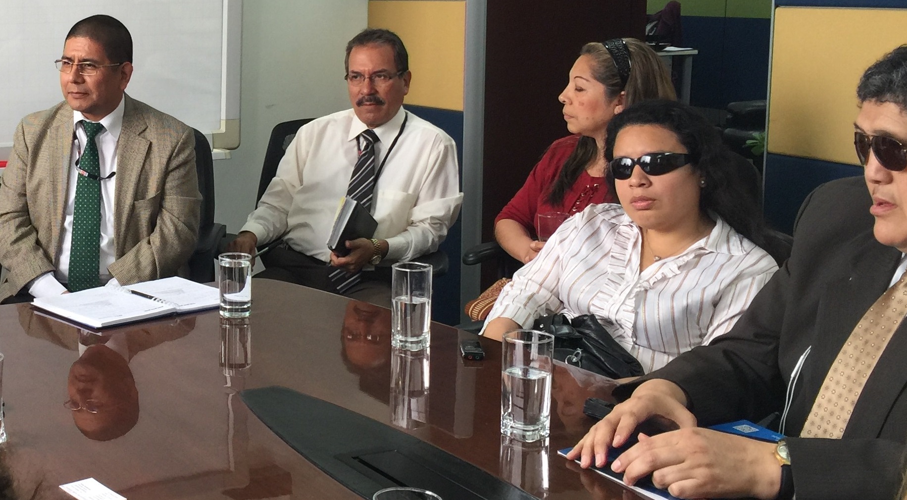 A group of people, some with and some without disabilities, sit at a table in a conference room listening to an unpictured speaker. They are all wearing business clothing.