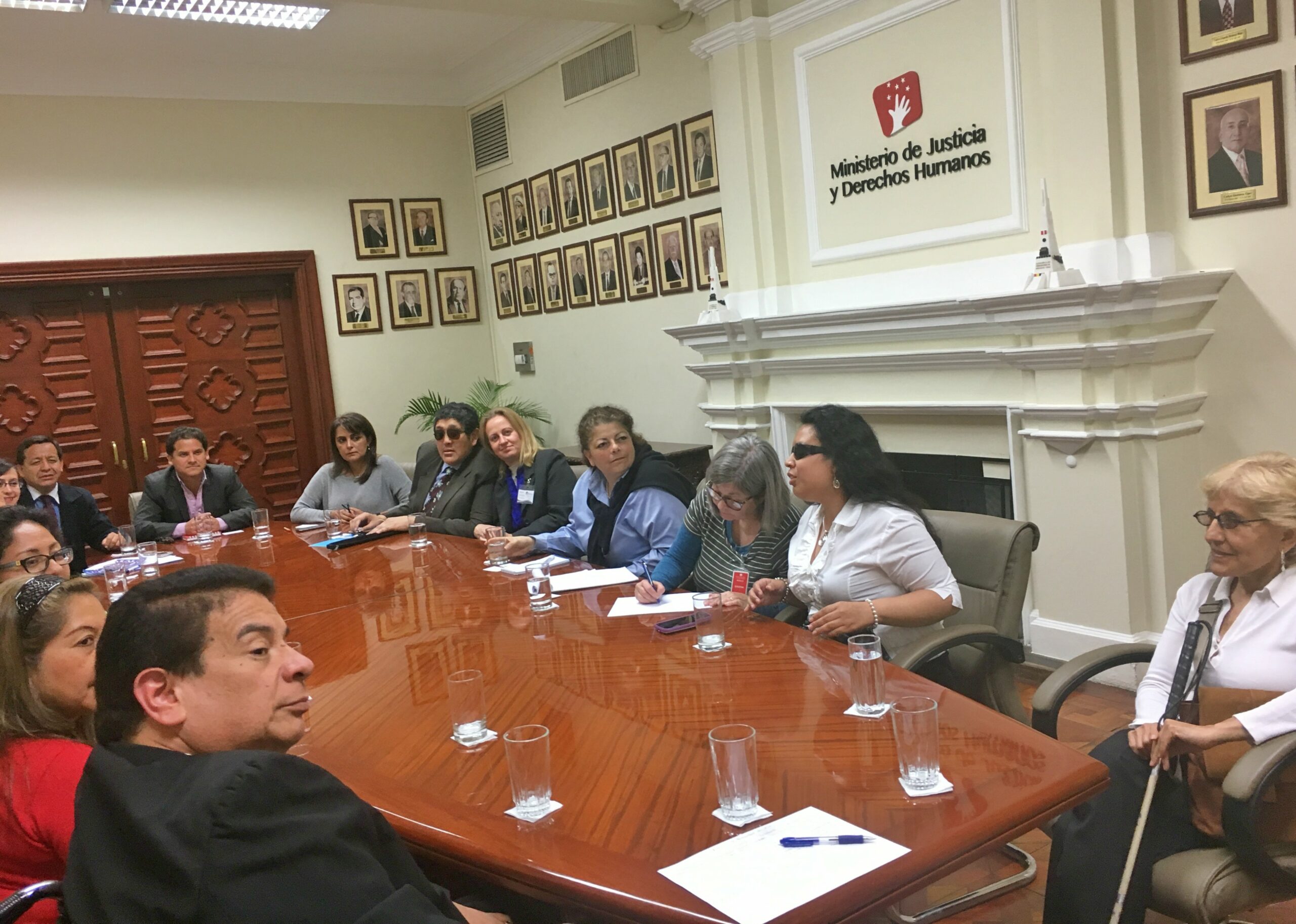 A group of people, some with and some without disabilities, sit around a large conference table. A sign on the wall reads in Spanish "Department of Justice and Human Rights."