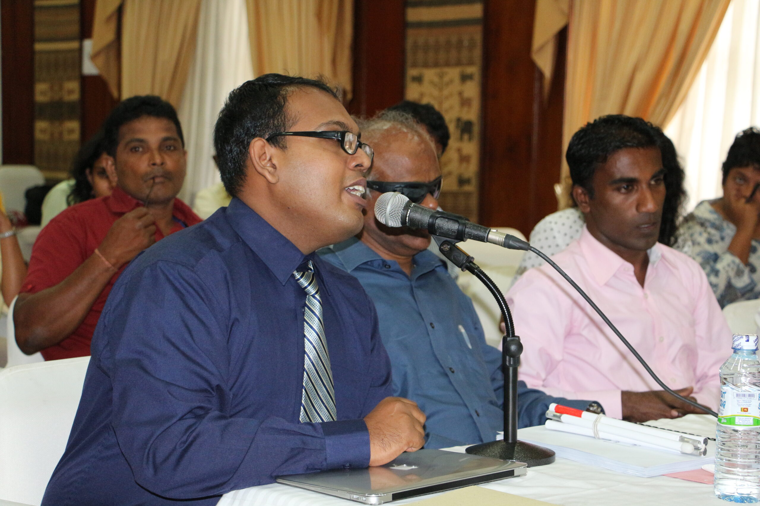 A man wearing professional clothing speaks into a microphone. He is surrounded by several other men sitting in the room to the side and behind him.