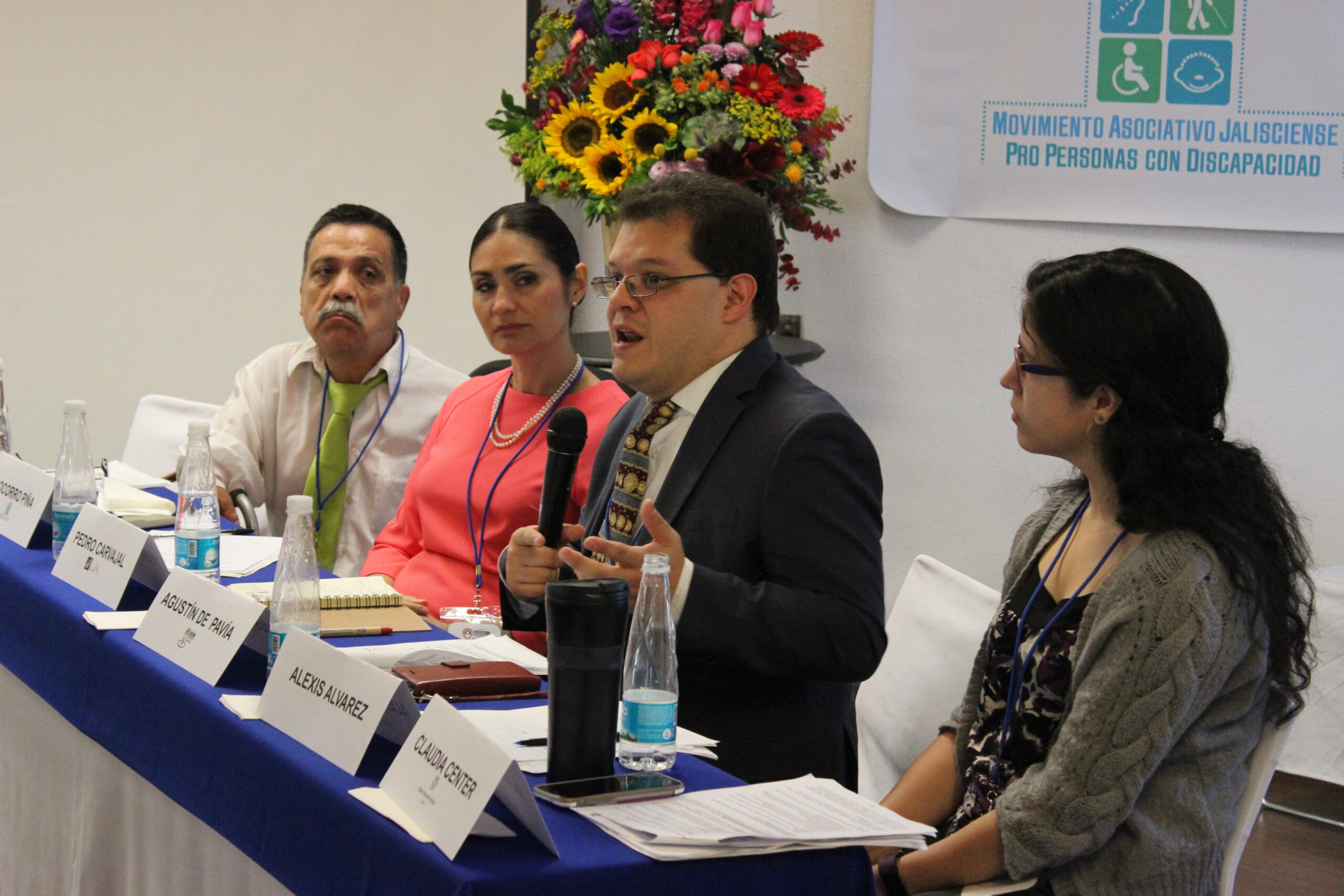 Four panelists sit at a table while one speaks using a microphone. The MADIJAL logo is posted on the wall behind